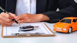 Homem mostrando apenas as mãos, assinando algo em uma folha, à sua frente há uma chave de carro e um carro em miniatura laranja.